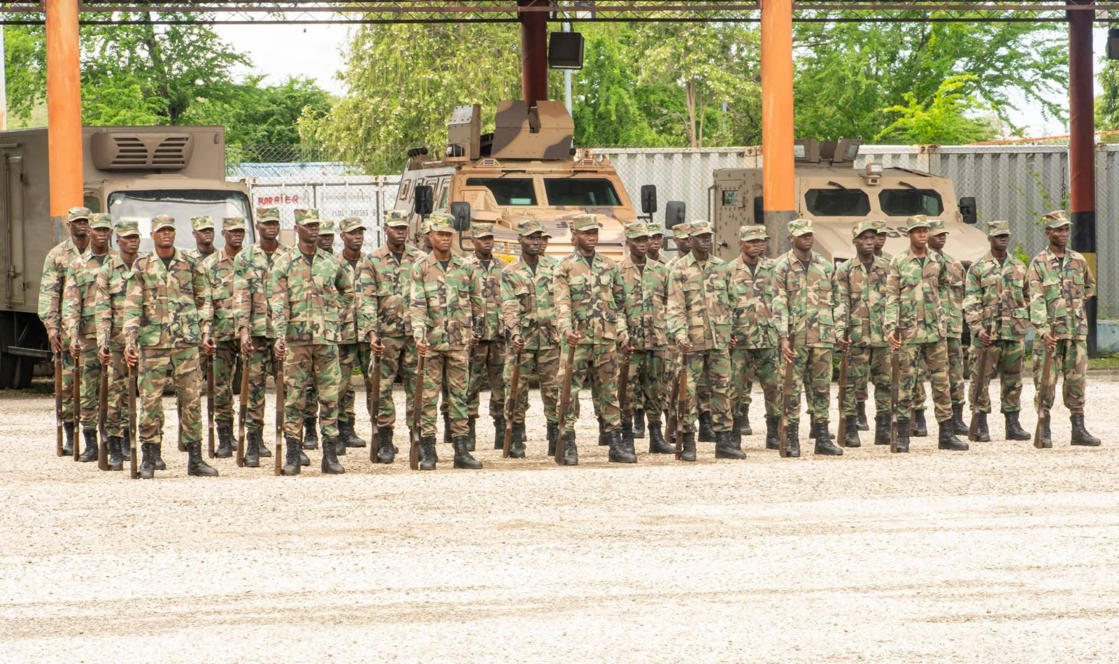 Line of soldiers in camouflage uniforms with rifles standing at attention in a military yard with armored vehicles behind them.