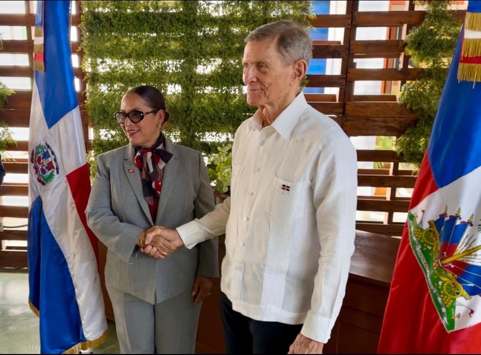 Two formally dressed individuals shake hands at a ceremony, with national flags in the background.