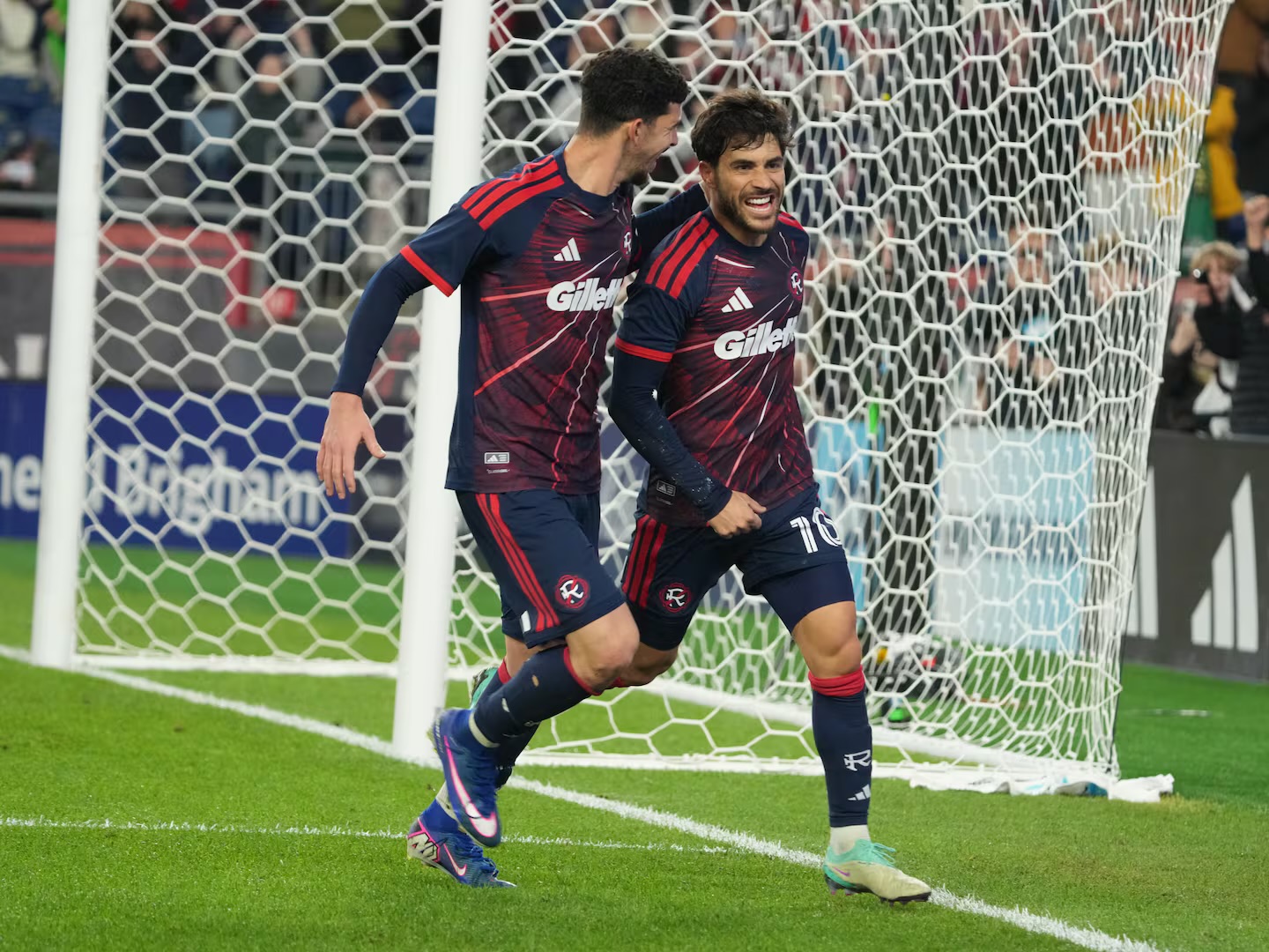 Two soccer players in maroon jerseys celebrate near the goal net on a green field, after a score.