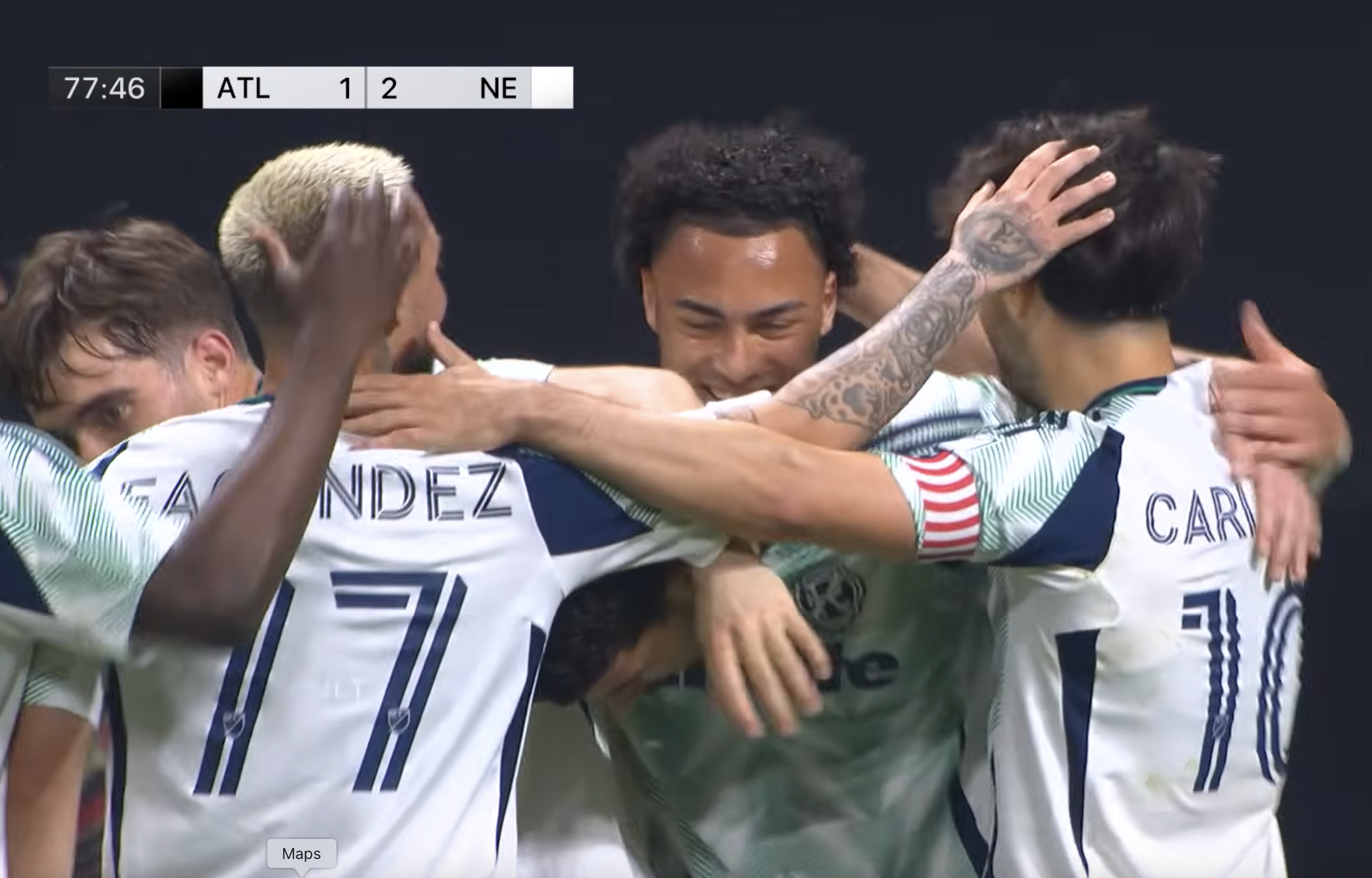 Soccer players in white jerseys form a group hug, celebrating a goal or win on the field.