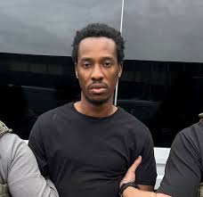 Man with short curly hair in a black T-shirt stands in front of a dark background, looking at the camera.