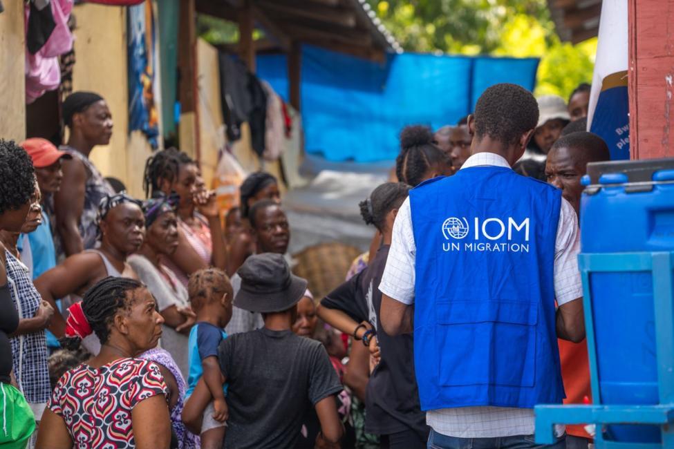 Aid worker in a blue IOM UN Migration vest speaking with a crowded group outdoors.