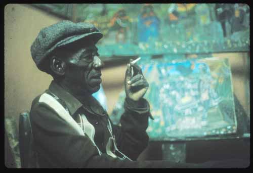 Andre Pierre - Caribbean Television Network Elderly man in a flat cap sits in a painter's studio, examining a lit cigarette between his fingers with colorful canvas paintings in the background.
