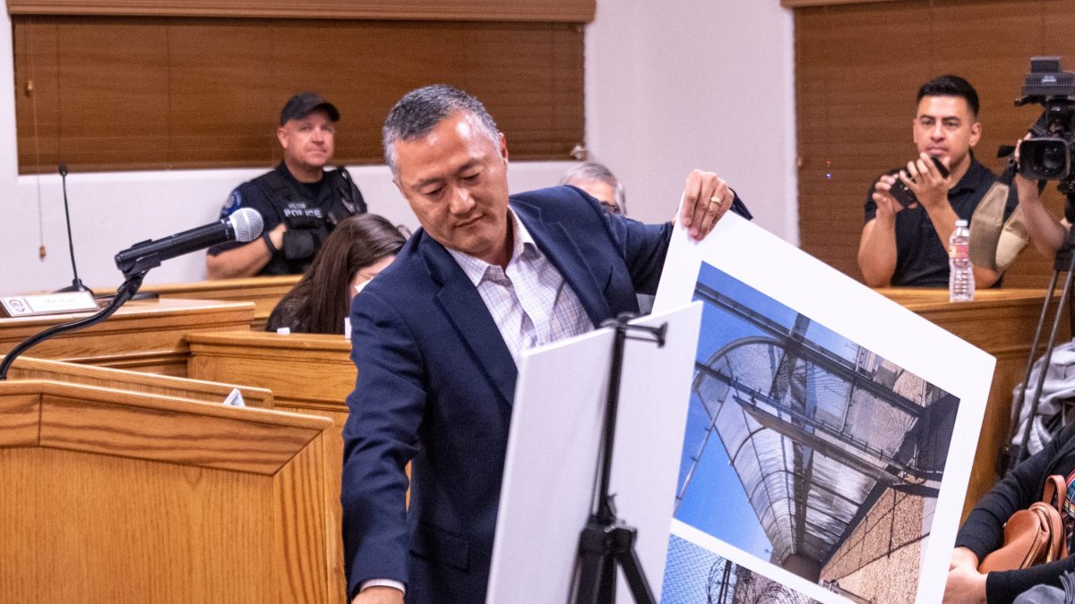 Man in a suit presents large photograph prints on a stand in a courtroom, with a police officer and media in the background.