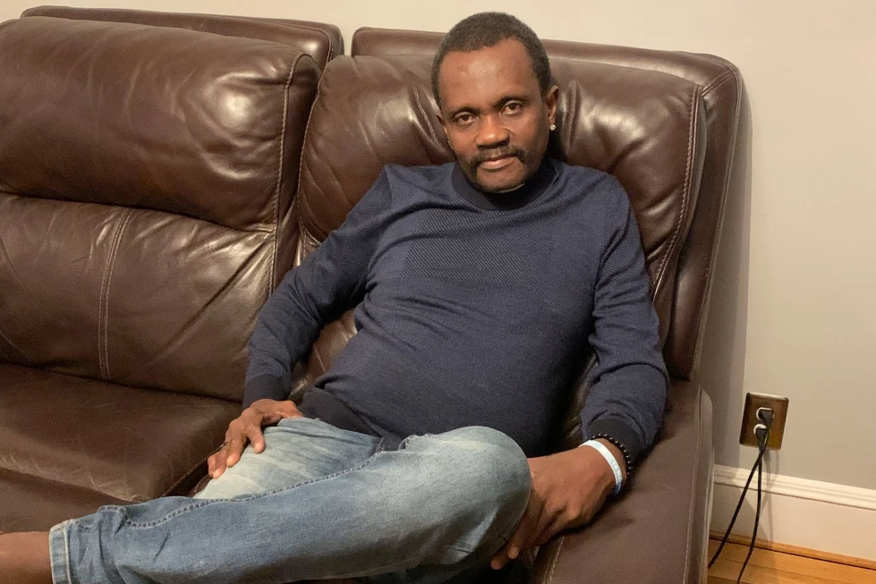 Man seated sideways on a dark brown leather sofa, wearing a navy blue sweater and light blue jeans, with one leg crossed and a relaxed expression in a room with an outlet and cable nearby.