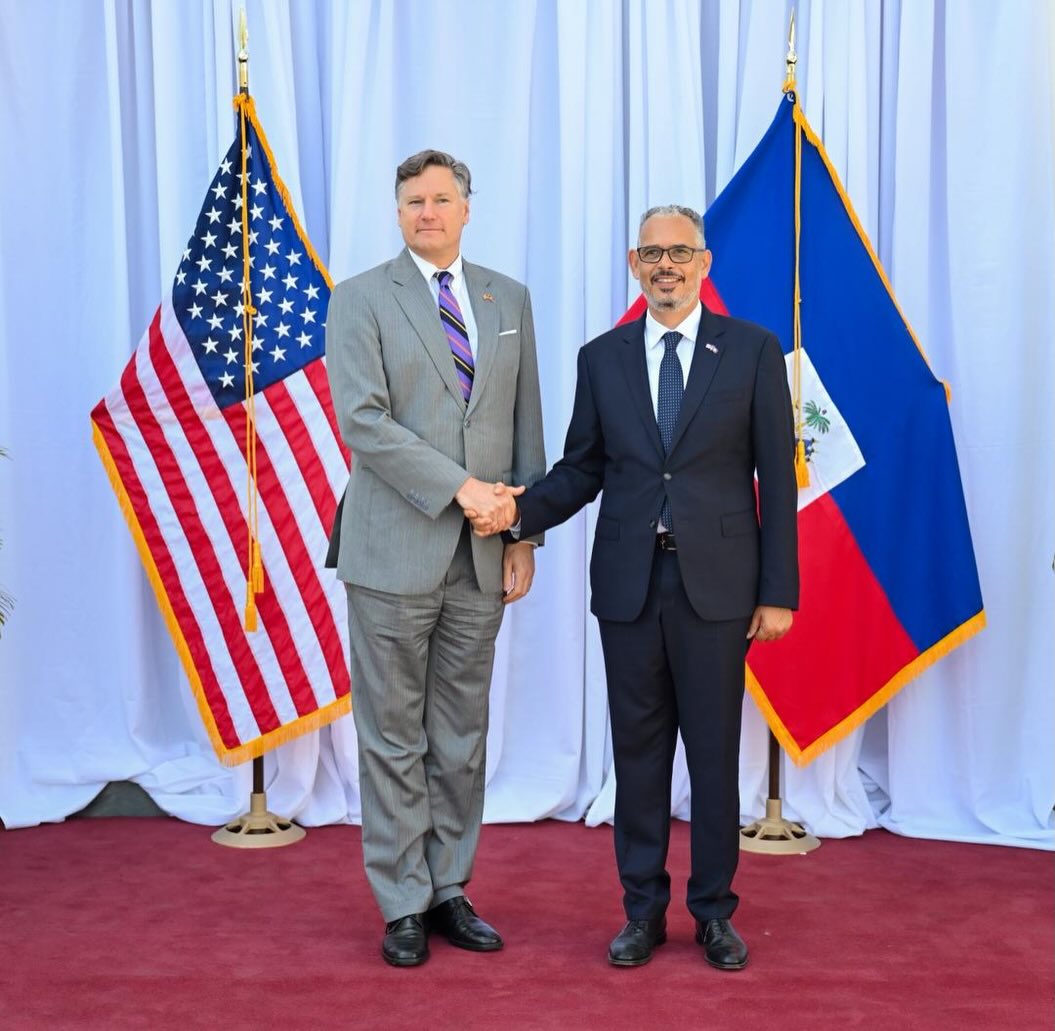 Two men in suits shake hands in front of flags—the U.S. flag on the left and another national flag on the right—standing on a red carpet against white drapes.