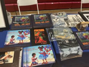 IMG_0083 - Caribbean Television Network Stacks of colorful art books, including 'Who We Art' and children's drawings, laid out on a blue table at a display booth.