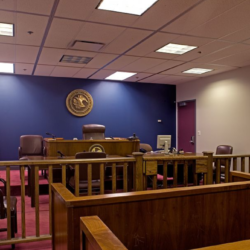 Empty courtroom with a raised judge's bench, wooden railing, and a seal on the blue wall behind it.