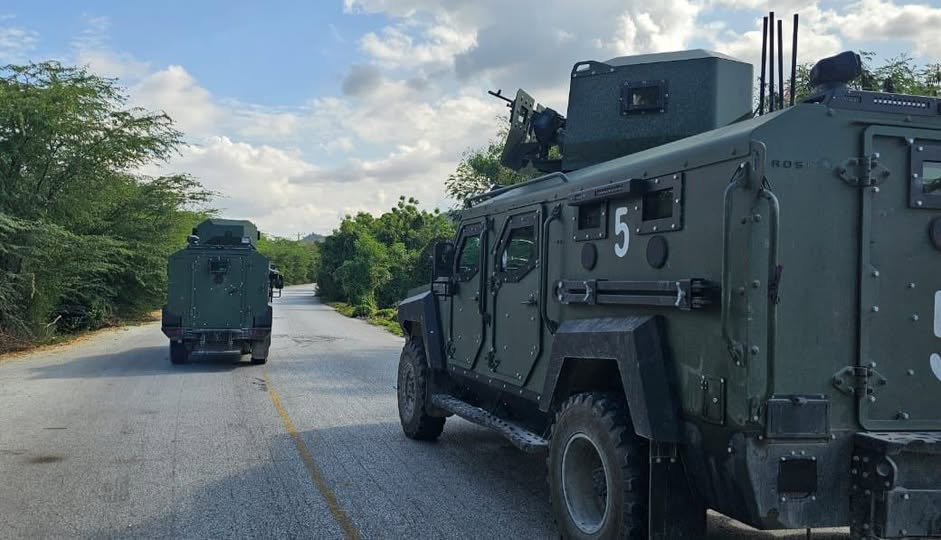 Armored military vehicle with the number 5 on a rural road, another vehicle in the distance, trees along the roadside.