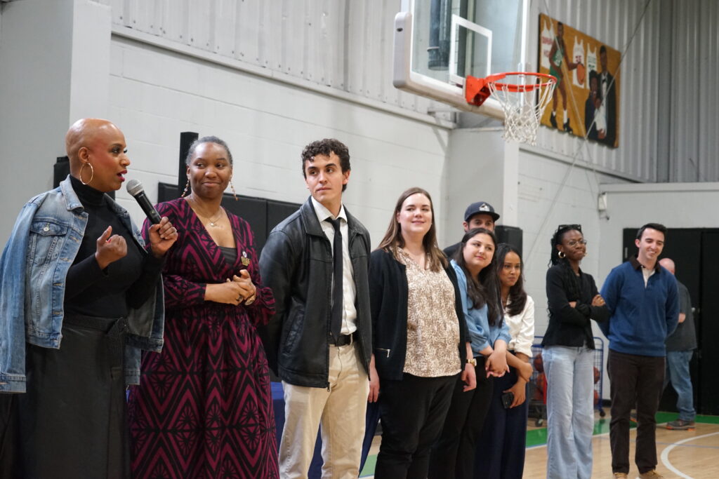 Diverse group of people standing in a gymnasium, with a woman speaking into a microphone on the left.