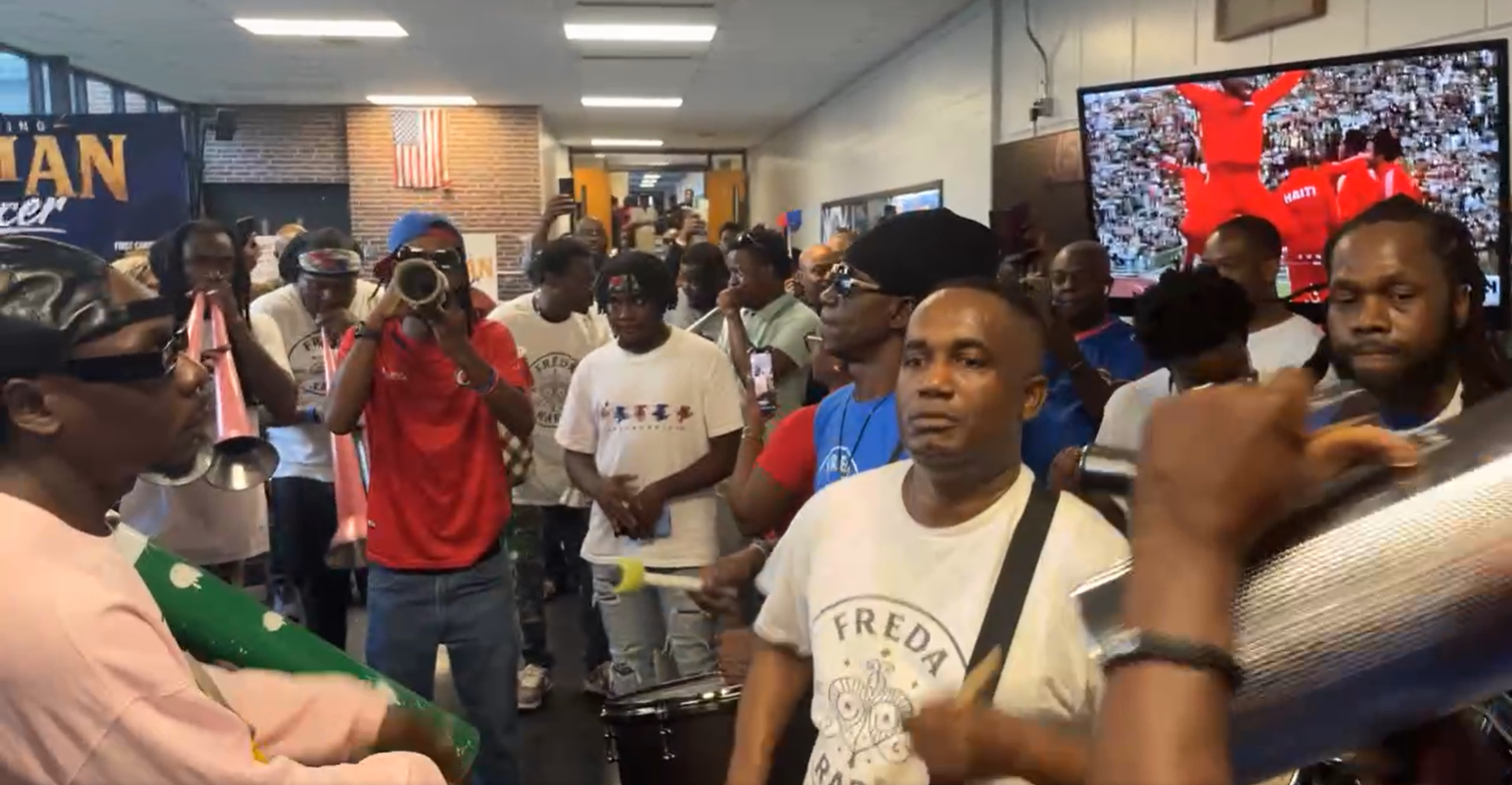 Rara Frantzdy - Caribbean Television Network Crowded indoor community event with musicians and spectators, a drummer in foreground and a person filming with a camera.