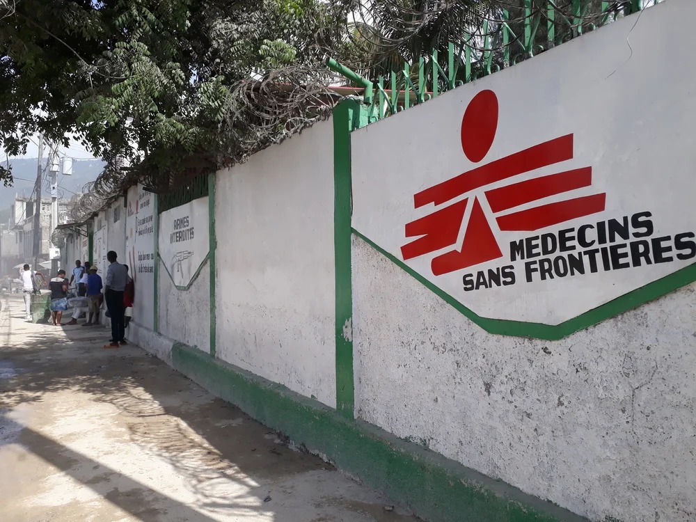 MSF wall mural with red emblem and text 'Médecins Sans Frontières' along a green-trimmed wall beside a dusty street with people nearby.