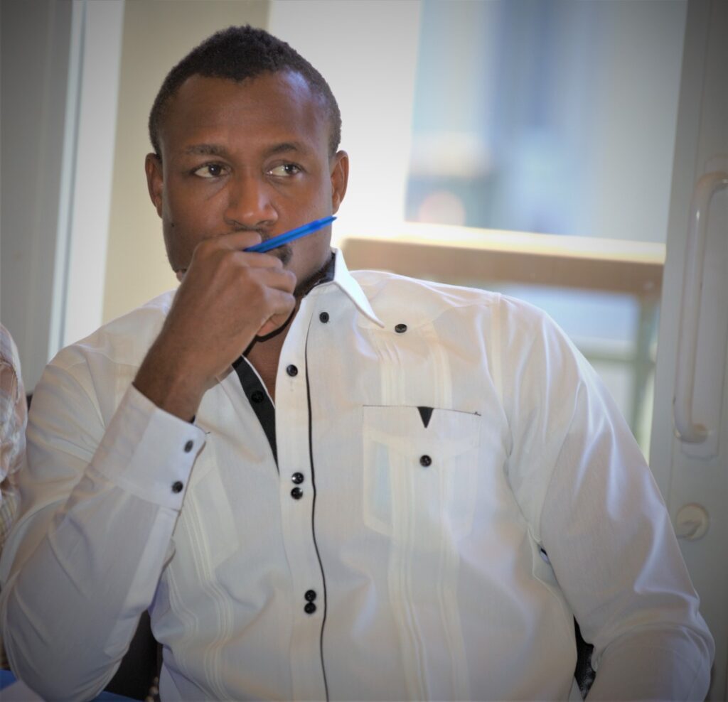 Man in a white shirt sits at a desk, holding a blue pen to his mouth in a thoughtful pose.