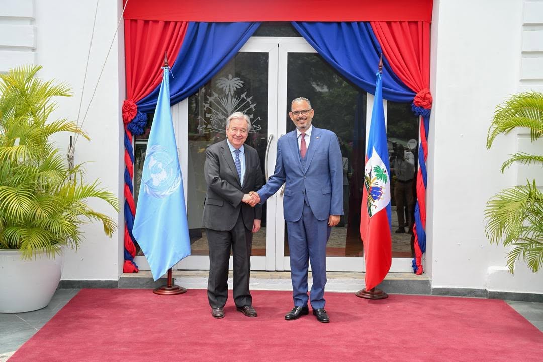 Two men in suits shake hands on a red carpet in front of a doorway framed by red and blue drapes, with a United Nations flag and a Haitian flag nearby.