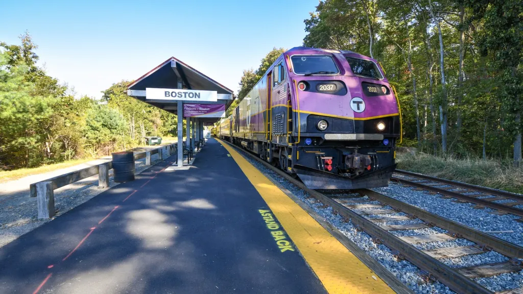 Purple diesel locomotive on a small station platform with 'Boston' sign, yellow safety edge, and trees in the background to the right.