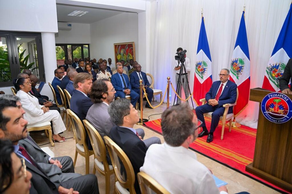 Formal Haiti event: a man in a blue suit sits on a stage beside three Haitian flags; audience in suits watches as a cameraman films.