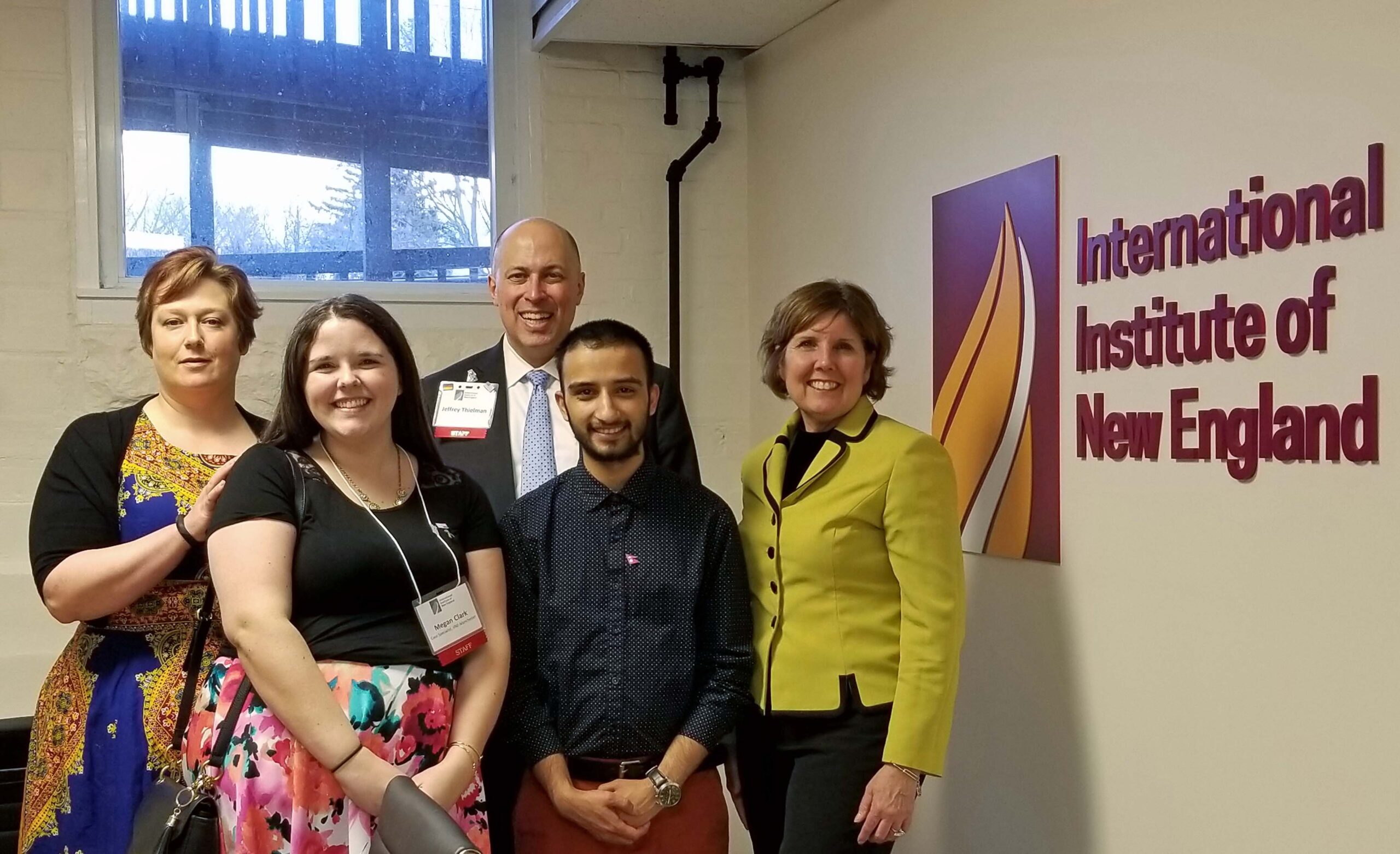 Five professionals pose for a photo in a hallway beside a large banner that reads 'International Institute of New England'.