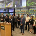Group of officials and first responders stand at a podium for a press briefing, with a wall of traffic-monitoring screens behind them showing maps and road footage.