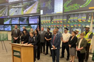 Group of officials and first responders stand at a podium for a press briefing, with a wall of traffic-monitoring screens behind them showing maps and road footage.