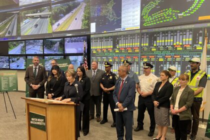 Group of officials and first responders stand at a podium for a press briefing, with a wall of traffic-monitoring screens behind them showing maps and road footage.