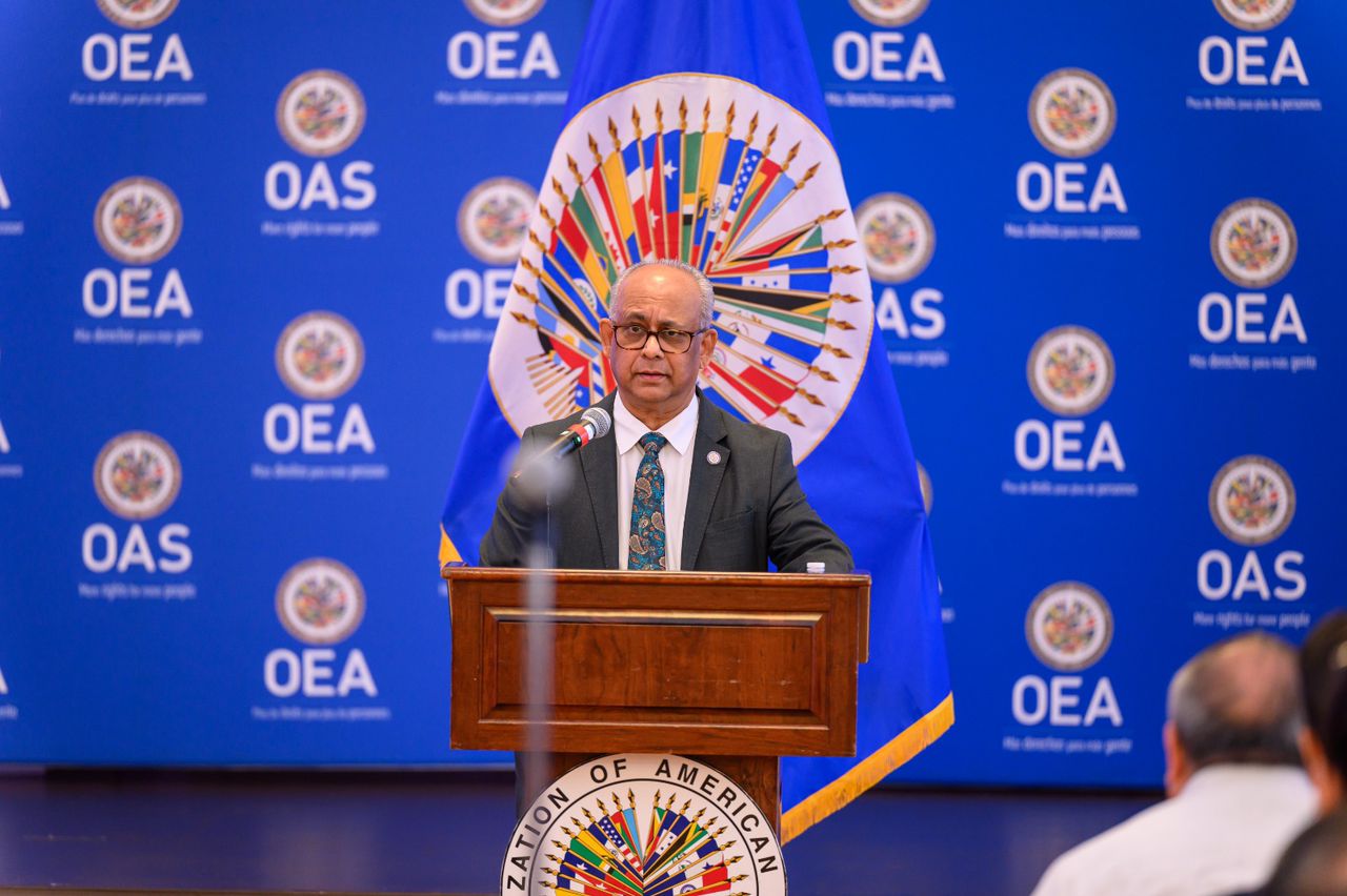 Male speaker in a suit delivers a speech at a podium with the OEA backdrop behind him and a blue flag nearby.