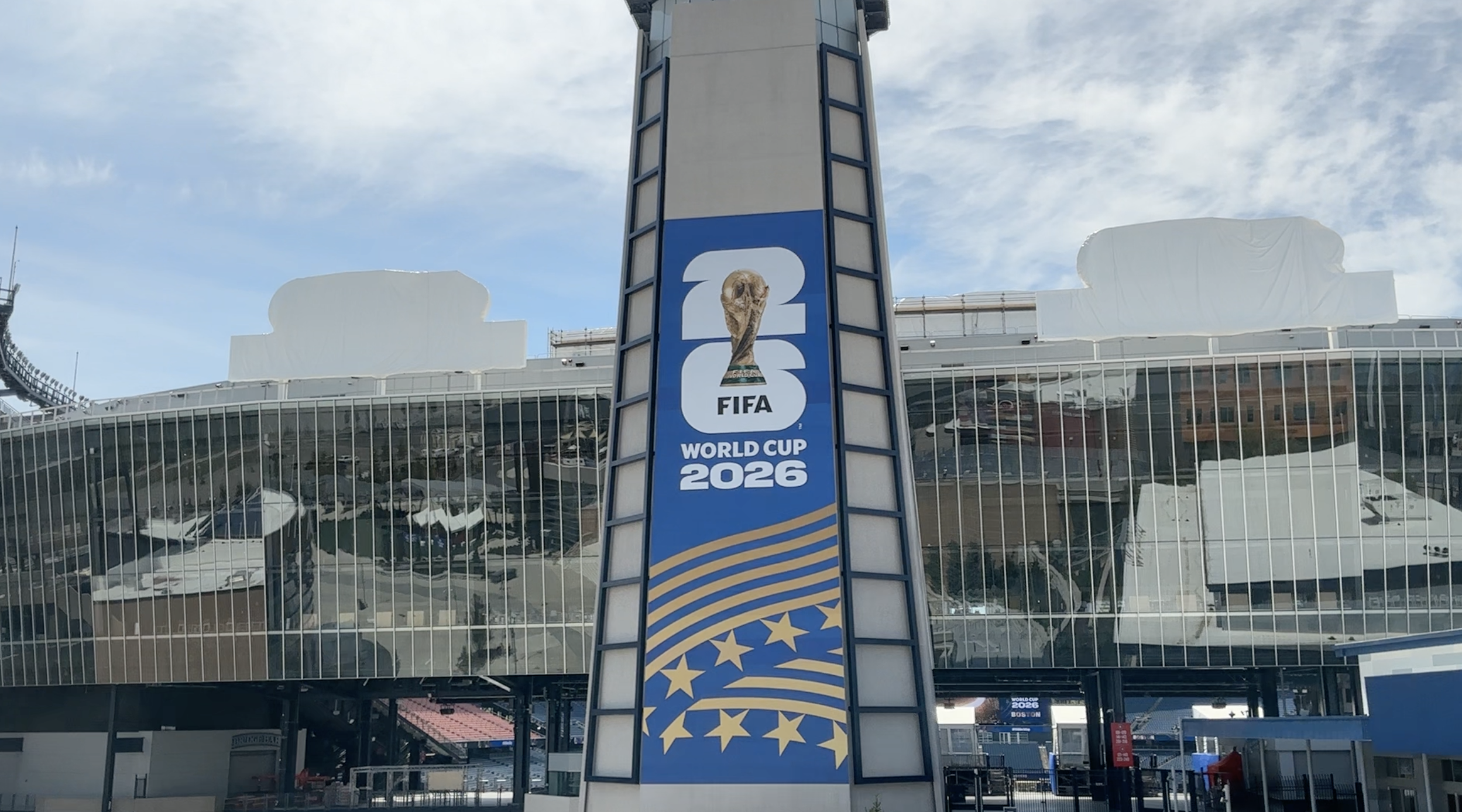 Vertical FIFA World Cup 2026 banner on a blue tower in front of a glass-front stadium, featuring the trophy image and waves motif.