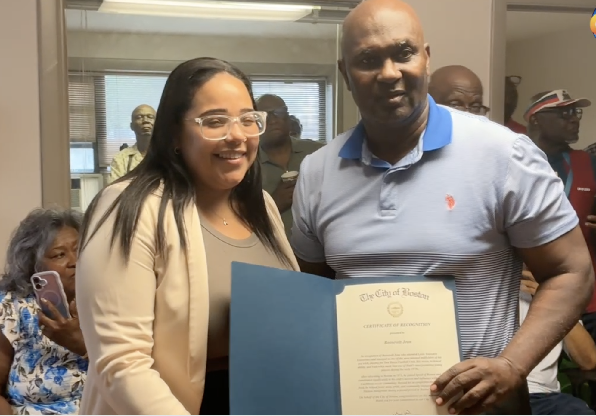 Screenshot 2026-06-11 at 85141 AM - Caribbean Television Network Smiling woman with clear glasses and beige blazer beside a man in a blue polo shirt holding a certificate at an award event, with others in the background.