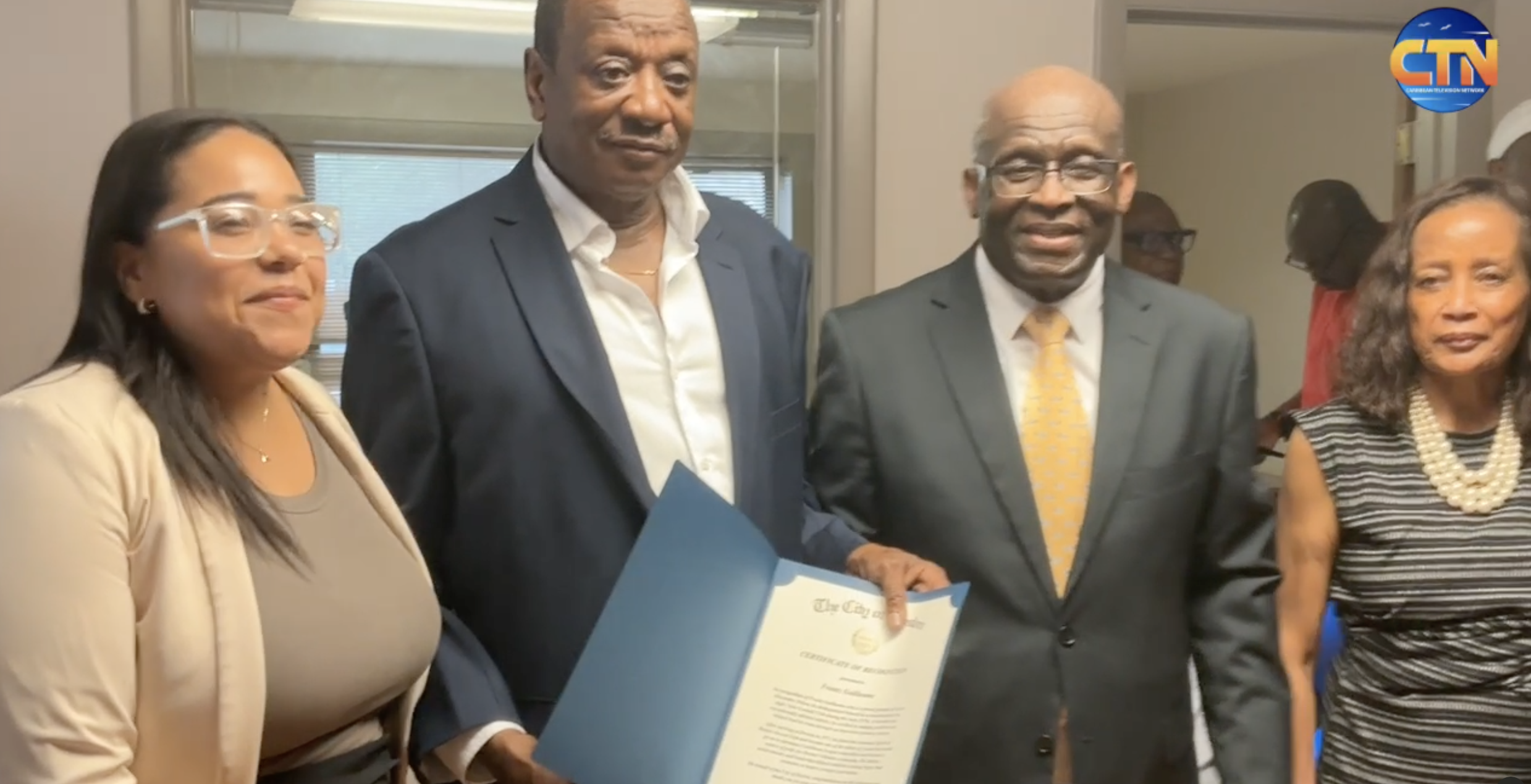 - Caribbean Television Network Four professionals in an office posing for a photo, one man holding a certificate in a blue folder, CTN logo in the top-right corner.