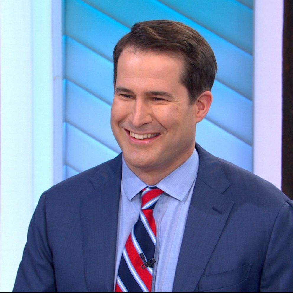 Smiling man in a blue suit and striped red, white, and blue tie on a television set.
