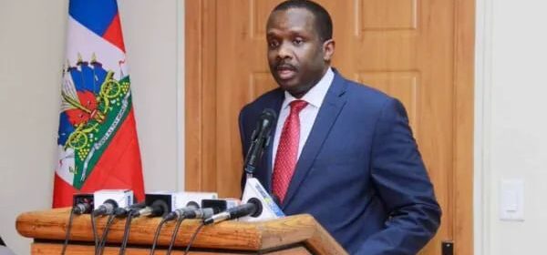 Man in a navy suit and red tie speaks at a podium with microphones during a press conference, Haitian flag visible in the background.