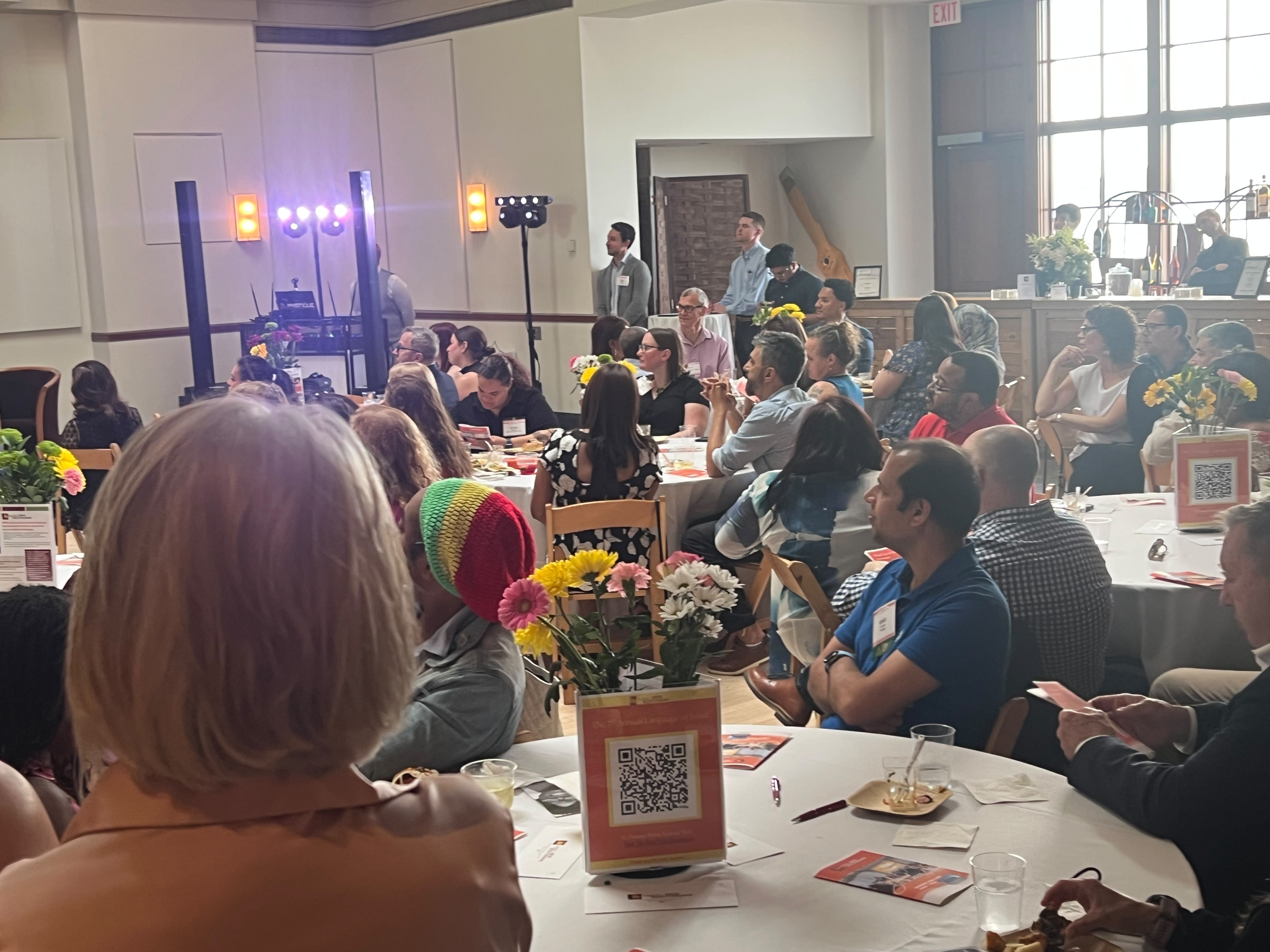Crowded banquet hall with round tables, attendees eating and chatting, colorful flower centerpieces, and a DJ area with stage lights in the background.