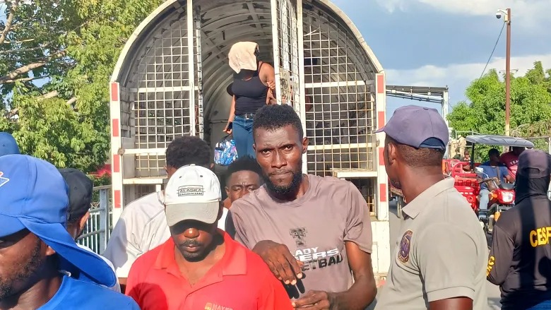 People disembarking from a large cargo truck with barred rear, several men in casual clothing and caps on a sunny street.