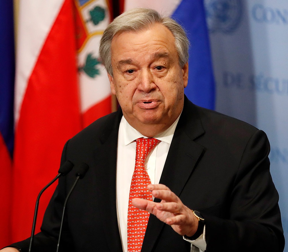 An older man in a dark suit and red tie speaks at a podium with two microphones, gesturing with his hand, flag backdrop behind him.