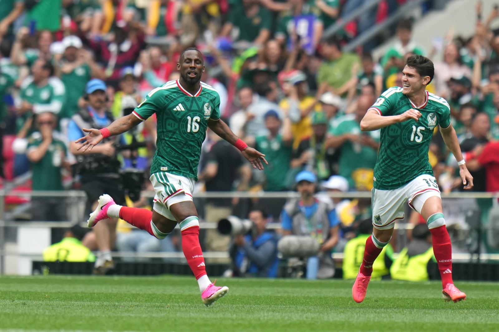 Two Mexican soccer players in green jerseys celebrate while running on a sunny field as the crowd cheers in the stands behind them.