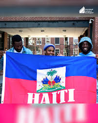 images - Caribbean Television Network Three people smiling while holding a Haiti flag with blue and red stripes and a central coat of arms in front of a brick building.