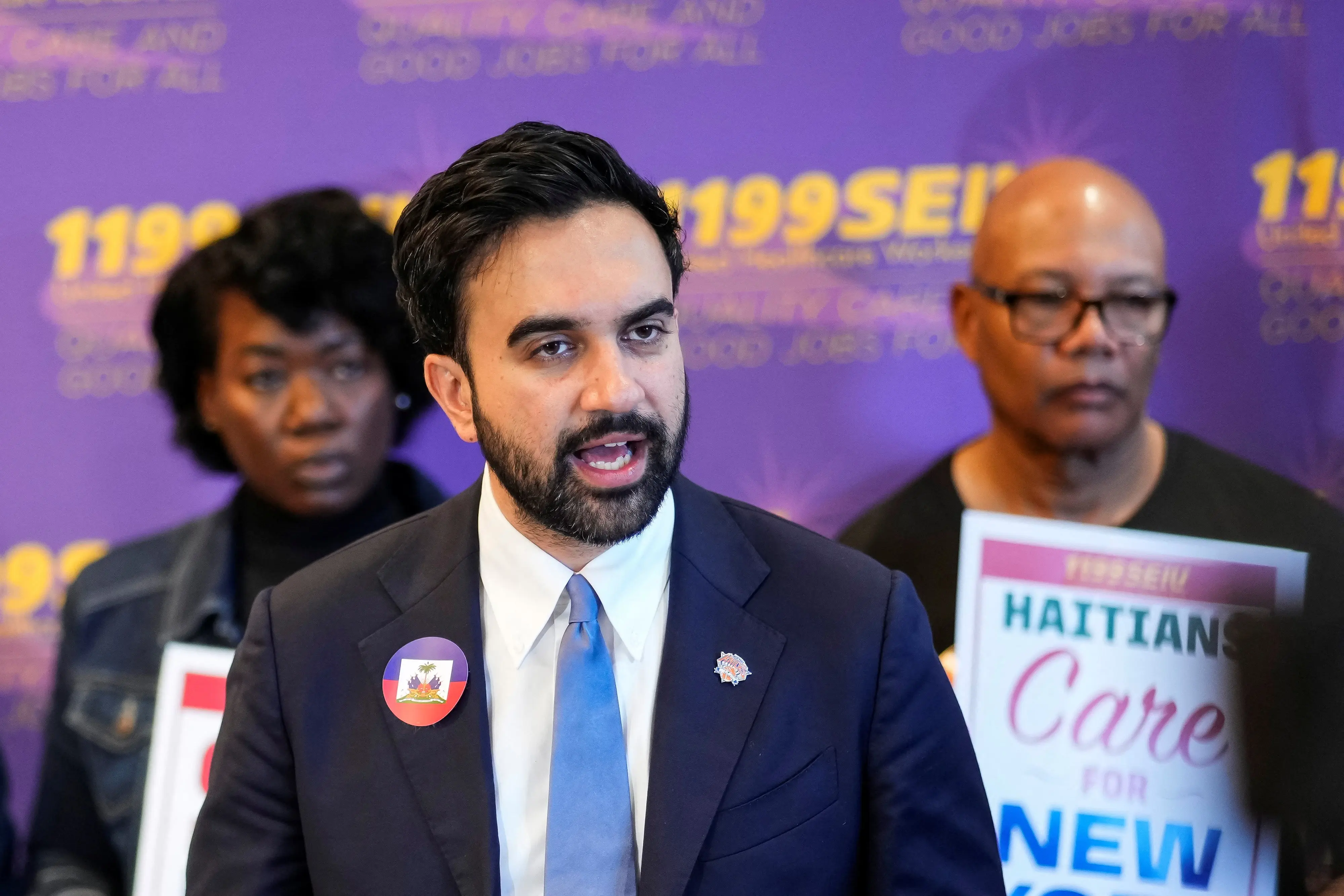 Man in a dark suit and blue tie speaks at a press conference, purple backdrop behind him