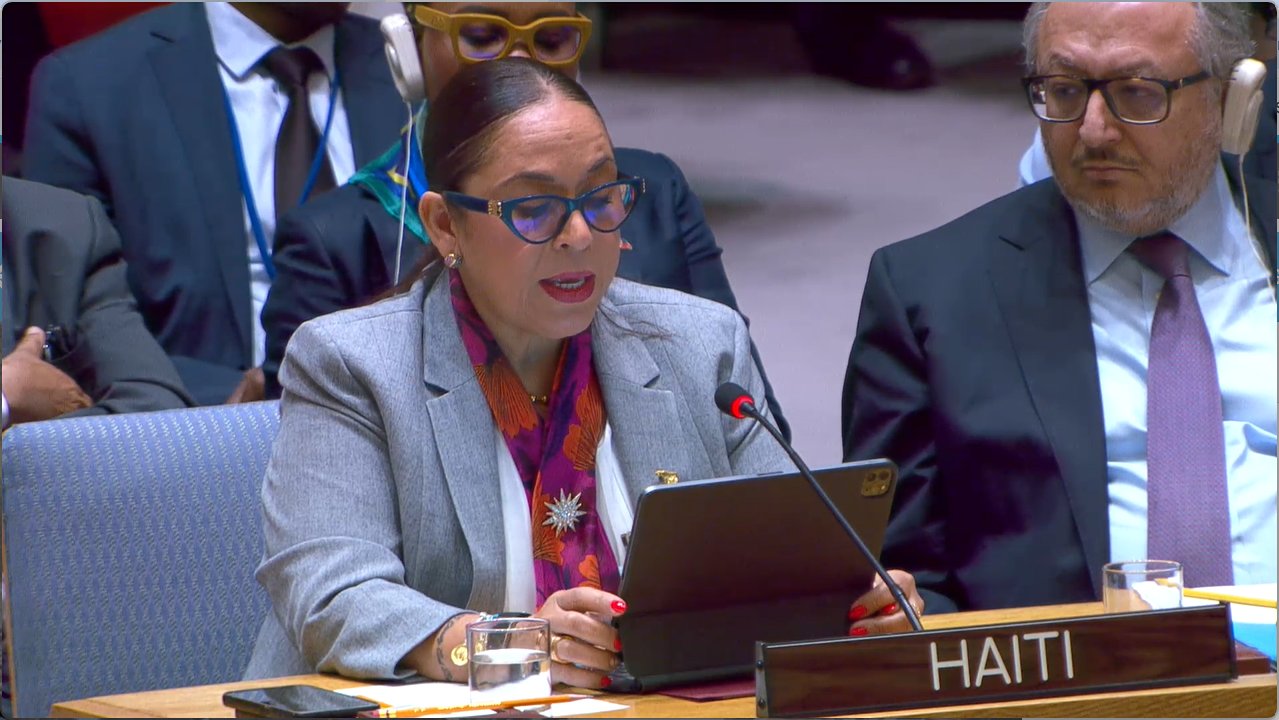 Haitian delegate speaks at a formal meeting, reading from a tablet, wearing glasses and a gray blazer, with a microphone in front and 'HAITI' nameplate visible.