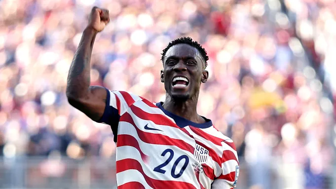 Soccer player wearing a USA 20 jersey celebrates with a raised fist on the field at a crowded stadium.