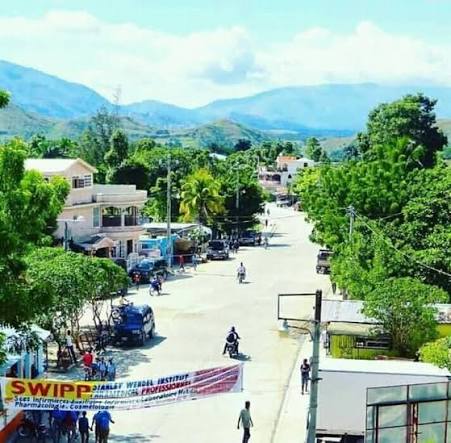 Sunny tropical street scene with low buildings, green trees, and motorbikes along a wide road; hills in the distance and a banner across the street.