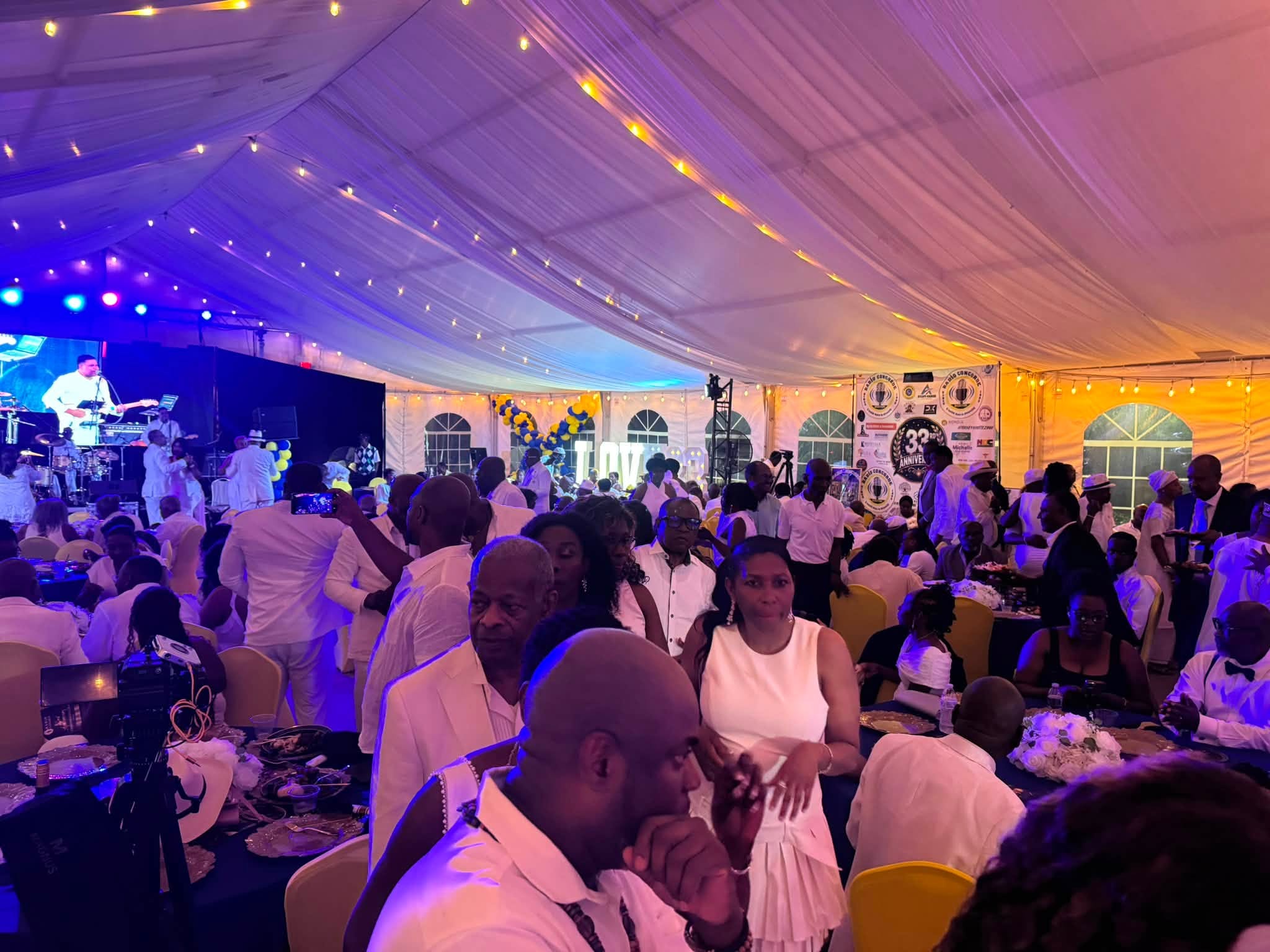 A crowded gala under a white tent with stage musicians illuminated by blue and purple lights and string bulbs overhead.