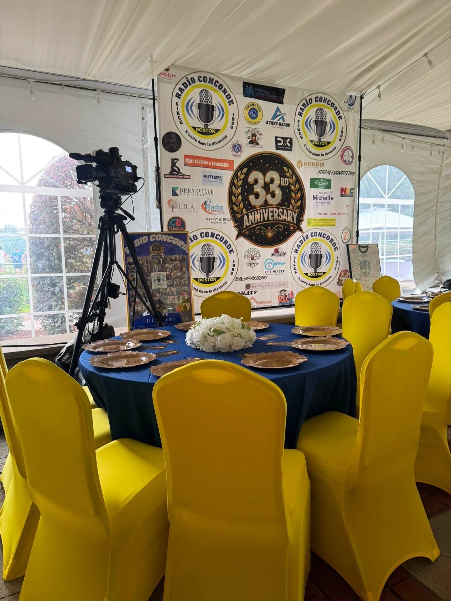 Concorde celebration 33 ans - Caribbean Television Network Event setup inside a white tent with a wall of sponsor banners, a camera on a tripod, round blue-dusted tables, and bright yellow chairs.