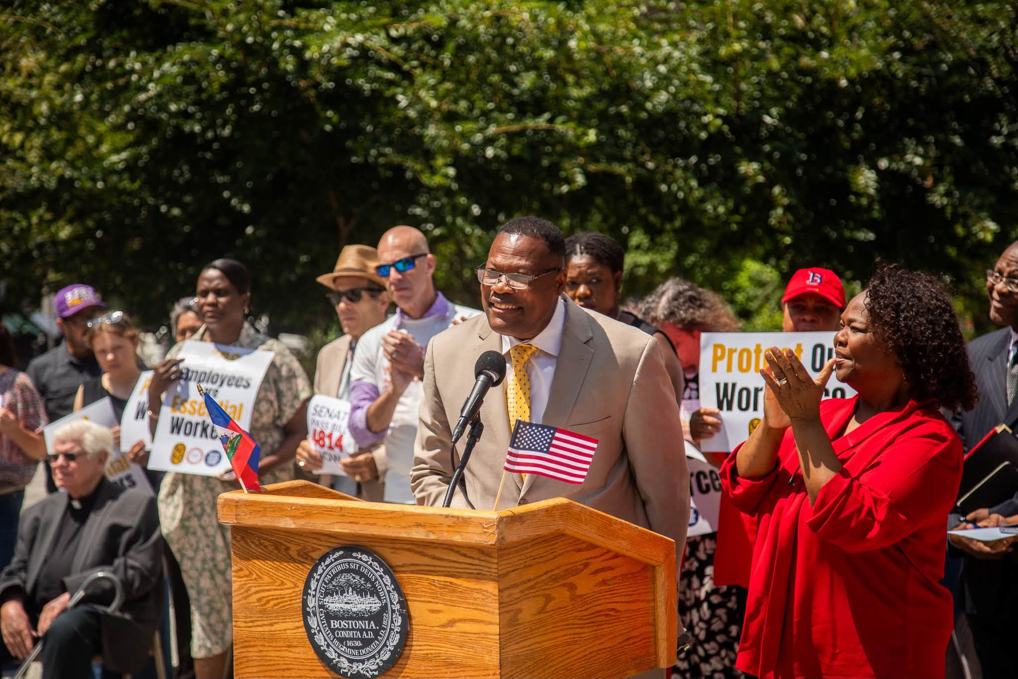 Dieufort Fleurissaint - Caribbean Television Network Man in a tan suit speaks at a wooden podium with an American flag, surrounded by protesters holding signs and banners.",