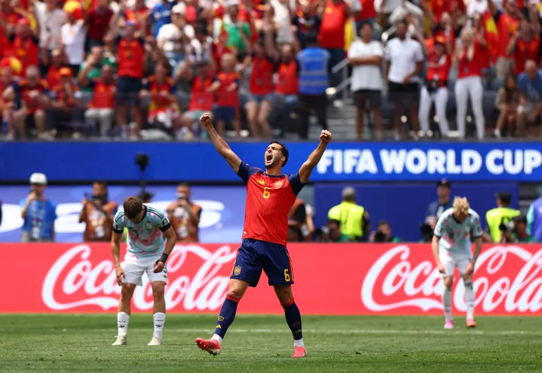 France vs Spain - Caribbean Television Network Soccer player in red and blue jersey (#6) raises arms in celebration on a green pitch with fans behind and Coca-Cola ads nearby