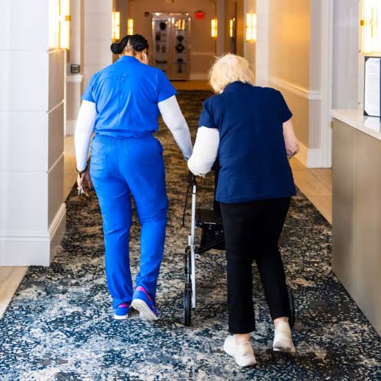 Caregiver in blue scrubs helping an elderly woman walk with a walker down a hospital corridor with bright walls and carpeted floor.