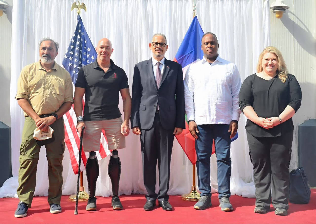 Five adults stand side by side on a red carpet in front of flags, posing for a formal group photo; the second person from the left has prosthetic legs visible beneath shorts still wearing black athletic shoes.