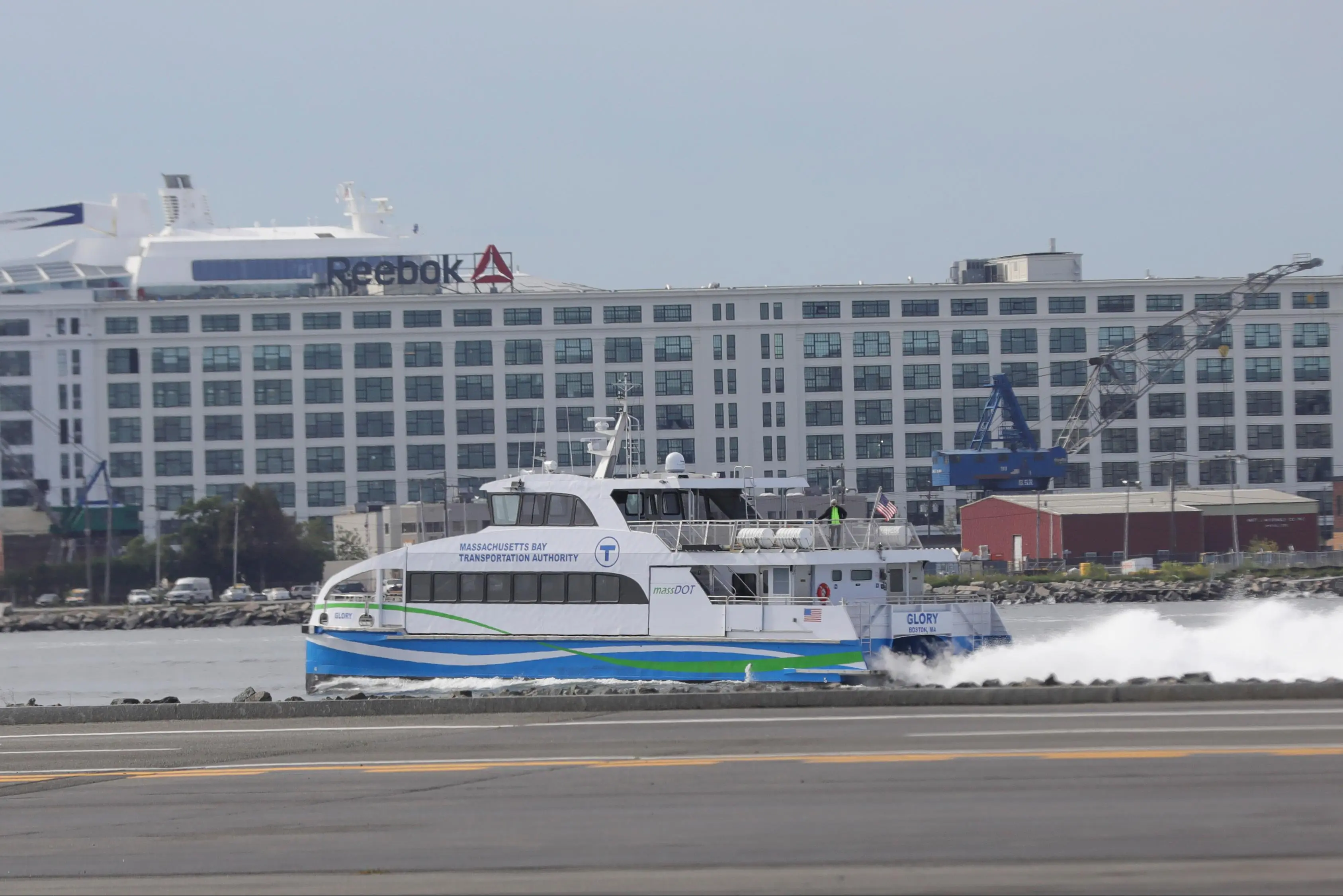 White waterfront building with a prominent Reebok logo overlooking a harbor; in the foreground, a MA Bay Transportation Authority ferry named 'Glory' on the water.