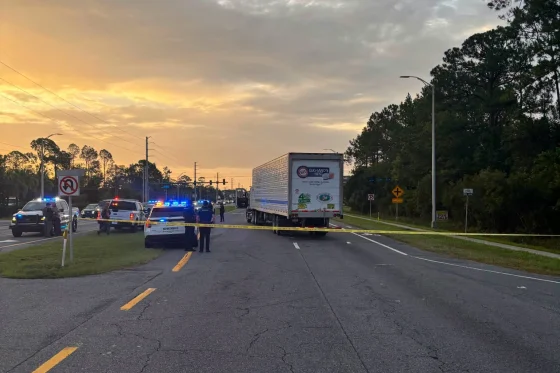 Roadway blocked by police and a large truck at sunset, with yellow tape across lanes and emergency lights flashing.