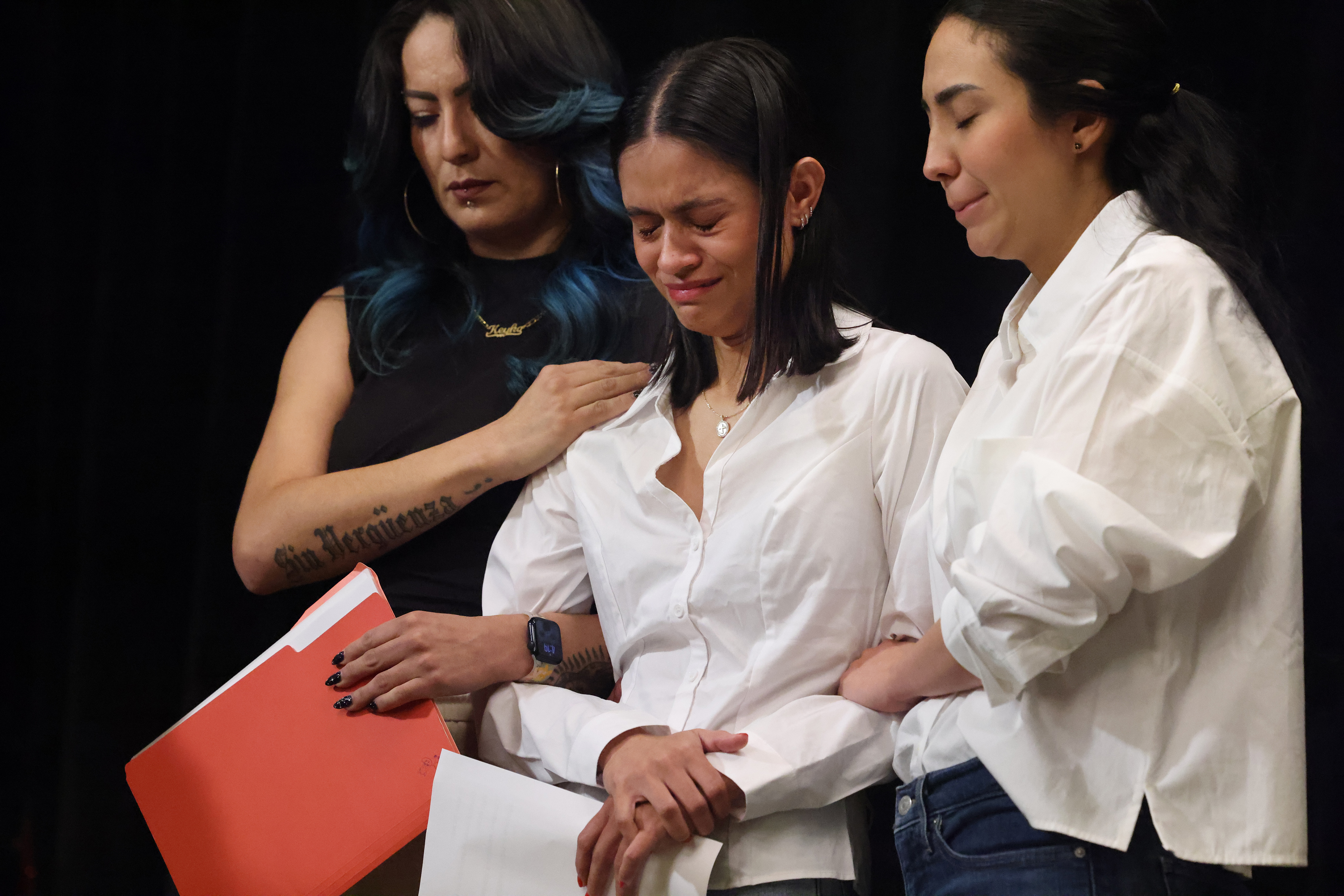 16presser - Caribbean Television Network Three women on a stage: a distressed woman in a white blouse is comforted by two others, one with blue-tinted hair in black, the other smiling, while they hold papers together.