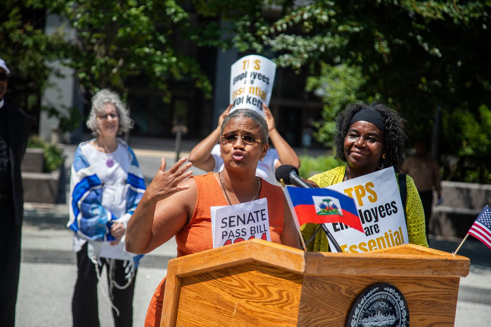Natacha Clerge2 - Caribbean Television Network Speaker at a protest podium, microphone in front, with signs demanding action like 'Senate Pass Bill' as people rally behind. An American flag is visible on the side.