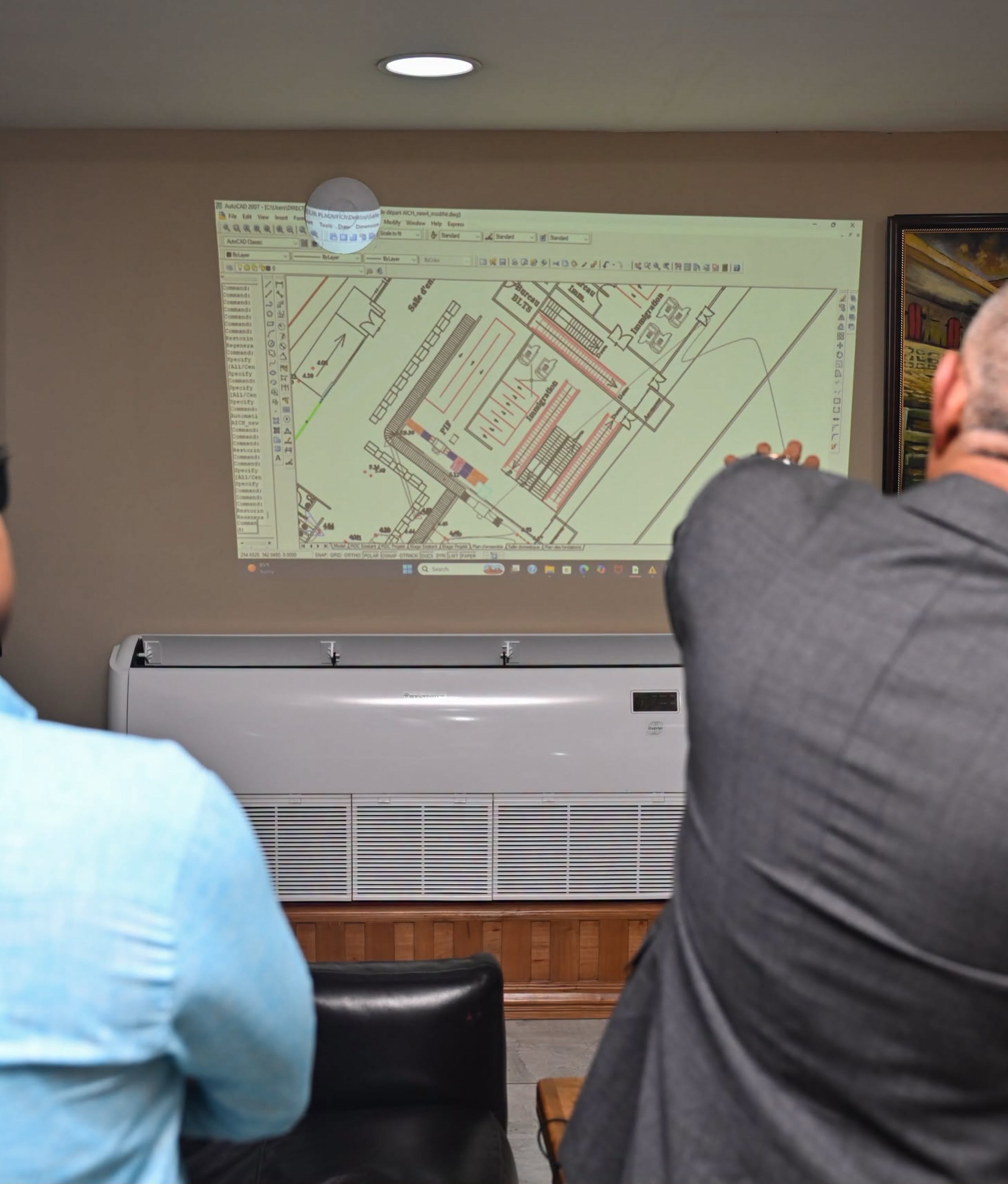 Two people sit facing a projected architectural floor plan on the wall in a meeting room.
